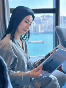 在Lady Hera 希拉月子中心客房內，剛生產完的媽媽坐在窗邊閱讀雜誌，窗外可見海港與城市天際線
